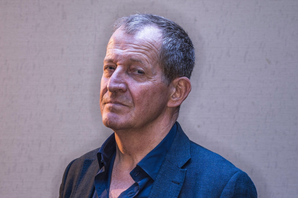 Close-up portrait of Alastair Campbell looking toward the camera with a thoughtful expression, wearing an unbuttoned blue shirt and navy blazer.